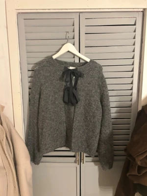 Grå stickad tröja med rosett från Vila - Mysig grå stickad tröja från Vila med lång ärm och en snygg svart rosett i halsringningen. Tröjan har en fluffig struktur och avslappnad passform, perfekt för kalla dagar. Enkel men ändå trendig design som passar till det mesta.