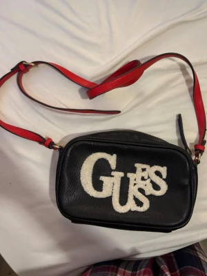 Svart axelväska från Guess - Snygg svart axelväska från Guess med vit logga i teddy på framsidan och röd axelrem. Väskan har guldfärgad dragkedja och ett mönstrat innerfoder med Guess-loggor. Perfekt storlek för det viktigaste och ger en cool touch till din outfit.