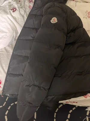 Svart Moncler dunjacka med logga - Säljer en svart dunjacka från Moncler med stor logga på ärmen och breda, quiltade partier. Jackan har dragkedja framtill och två sidofickor med dragkedja. Insidan har ett band med Moncler-tryck. Perfekt för kalla dagar.