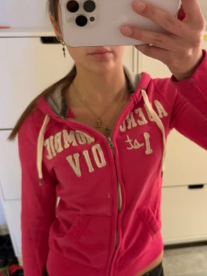 Rosa zip up hoodie Abercrombie & Fitch - Snygg rosa hoodie med dragkedja från Abercrombie & Fitch. Stor vit brodyrtext på bröstet, vita snören i huvan och fickor framtill. Mjuk insida och klassisk passform, perfekt för en avslappnad stil.