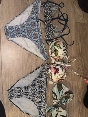 Fyra färgglada bikinidelar i olika mönster - Fyra olika bikinidelar: en blåvit trosa med blommigt mönster, en trekantsbikini med svartvitt zickzackmönster, en vadderad bikiniöverdel med tropiskt blommönster och en bandeauöverdel i grönt, brunt och blått abstrakt mönster. Perfekt för stranden eller poolen.
