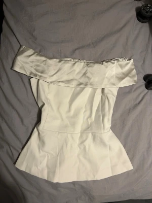 Vit offshoulder topp med volang - Trendig vit offshoulder topp med bred volangkant över axlarna och figurnära passform. Toppen har en lätt utsvängd nederdel och dragkedja i ryggen för enkel påklädning. Perfekt för dig som vill ha en stilren och modern look.