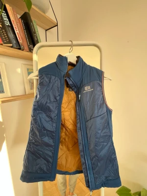 Blå dunväst från State of Elevate - Snygg blå dunväst från State of Elevate med guldfärgat innerfoder och hög krage. Västen har dragkedja framtill och diskret logga på bröstet. Perfekt för lager-på-lager och designad i Åre, Sverige. Passar dig som vill ha en sportig och stilren look. Nypris 2200kr