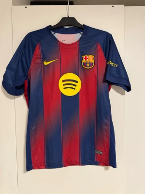 FC Barcelona matchtröja Lamine Yamal - Snygg FC Barcelona matchtröja i blått och rött med gula detaljer. Tryck med Lamine Yamal och nummer 10 på ryggen. Nike-logga, Spotify och UNHCR-tryck samt klubbmärke på bröstet. Korta ärmar och officiella Champions League-märken.