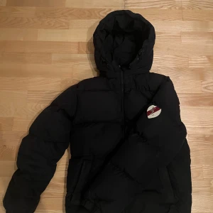 Svart pufferjacka från kingsland - Svart pufferjacka med huva och dragkedja framtill. Jackan har en broderad patch på ena ärmen och två fickor framtill. Perfekt för kyliga dagar och ger en cool streetwear-look. Superfint skick!! Priset kan diskuteras. 