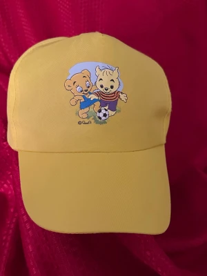 Gul keps med Bamse-motiv - Snygg gul keps med tryck av Bamse och Lille Skutt som spelar fotboll på framsidan. Kepsen har böjd skärm och justerbar rem baktill för perfekt passform. Tillverkad i mjukt tyg som känns bekvämt hela dagen.