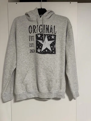 SCNDI Hoodie - Grå hoodie med tryck framtill där det står 'ORIGINAL FIT EST. 2024' och en stor stjärna omgiven av mindre stjärnor. Klassisk huva med dragsko och känguruficka. Perfekt för en avslappnad stil.