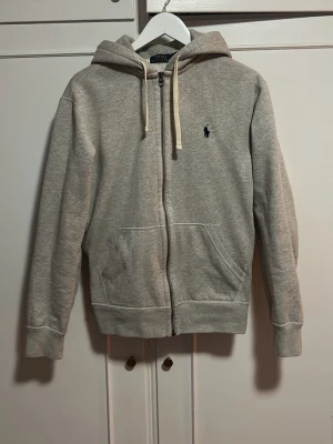 Grå zip hoodie från Polo Ralph Lauren - Snygg grå hoodie med dragkedja från Polo Ralph Lauren. Klassisk design med huva och vita snören, två fickor fram och den ikoniska lilla mörkblå loggan broderad på bröstet. Tillverkad i mjukt sweatshirtmaterial, perfekt för en chill och avslappnad stil.