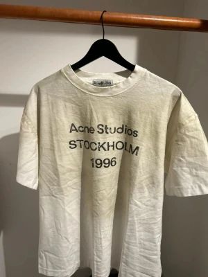 Vit Acne Studios t-shirt med tryck - Säljer en vit t-shirt från Acne Studios med svart tryck 'Acne Studios STOCKHOLM 1996' på framsidan. T-shirten har rund hals och korta ärmar, tillverkad i mjuk bomull. Perfekt för dig som gillar clean och stilren design med en touch av street.