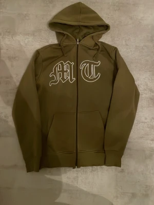 Tuttolente hoodie zip up - Olivgrön hoodie från tuttolente med dragkedja framtill och stora vita bokstäver 'M' och 'T' på bröstet. Klassisk huva och två fickor framtill. Tillverkad i mjukt material som passar perfekt för en avslappnad stil.
