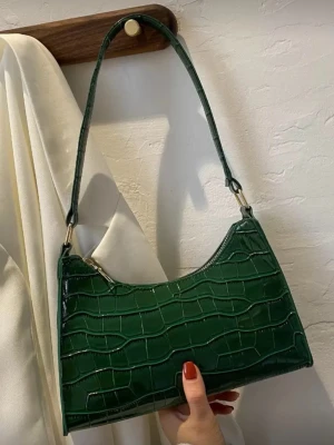 Grön croco handväska  - Trendig grön handväska i croco-mönstrat syntetmaterial med snygg glansig finish. Väskan har en böjd form och en smal axelrem i samma material. Stängs med dragkedja och har guldfärgade metalldetaljer. 