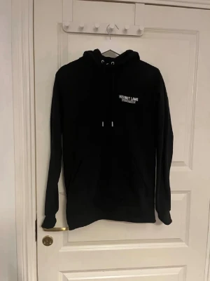 Svart hoodie från Helmut Lang XS - Svart hoodie från Helmut Lang med vit logga på bröstet. Klassisk modell med huva och dragsnören samt känguruficka framtill. Tillverkad i mjukt material som passar perfekt för en avslappnad stil.