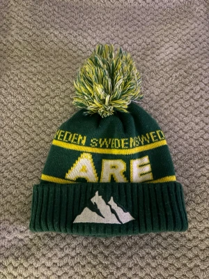 Grön Åre mössa med tofs - Grön stickad mössa med gul text 'SWEDEN' och 'ÅRE', samt vit bergslogga framtill. Mössan har en stor grön, gul och vit tofs på toppen och ribbad uppvikt kant. Perfekt för kalla dagar i fjällen. Inköpt i butiken i Åre. Limiterad färg så passa på! 