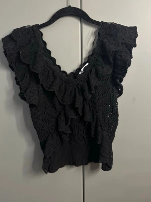 Svart virkad volangblus - Supersöt svart virkad blus med volanger och v-ringning. Toppen är ärmlös och har breda axelband med extra volangdetaljer. Perfekt för dig som vill ha en feminin och trendig look. Materialet känns luftigt och toppen är croppad i modellen.