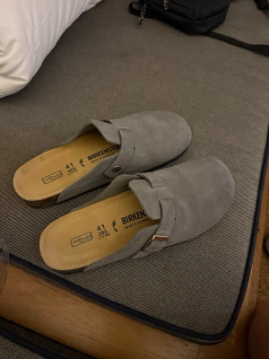 Gråa  Birkenstock tofflor - Säljer ett par gråa Birkenstock tofflor i mjuk mocka med klassisk rem och metallspänne. De har en öppen häl och rund tå, samt en bekväm innersula i naturfärgat material. Perfekta för chill dagar hemma eller ute.