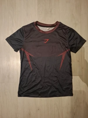 Svart röd compression shirt från Gymshark även kallad Gymshark Onix - Snygg svart sport-t-shirt från Gymshark med röda detaljer och logga på bröstet. Tillverkad i ett lätt och snabbtorkande material, perfekt för träning. T-shirten har rund halsringning och korta ärmar med en modern passform. Storleken är XXS men jag skulle säga att den passar S också efterssom det är så stretchigt material.