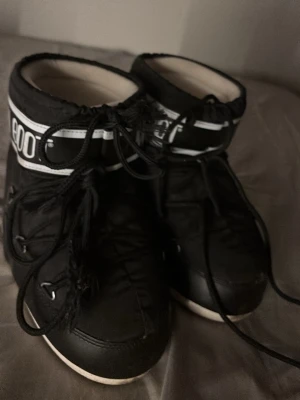 Svarta Moon Boot med snörning - Säljer ett par klassiska svarta Moon Boot med vit sula och tydlig logga runt skaftet. Bootsens ovandel är i syntetmaterial och de har snörning framtill för justerbar passform. Perfekta för kalla dagar och riktigt snygga till vinterlooken.OBS kmr tvätta dem noga innan jag skickar!