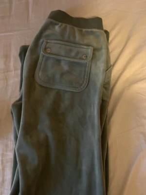 Gröna mjukisbyxor med fickdetalj - Mysiga gröna mjukisbyxor med bred mudd i midjan och en stor bakficka med lock och tryckknappar. Byxorna är uppsydda och sitter perfekt på mig som är 170 cm lång. Perfekta för chill dagar och har en skön loose fit. Materialet känns mjukt och fleece-liknande.