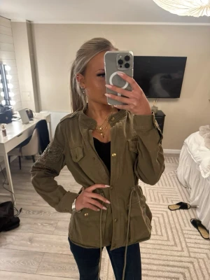 Olivgrön vindjacka med snörning - Snygg olivgrön vindjacka med snörning i midjan och flera fickor framtill. Jackan har guldiga knappar och patchdetaljer på ena ärmen. Perfekt för dig som vill ha en cool och avslappnad stil.