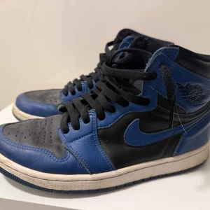 Nike Air Jordan 1 blå och svarta sneakers - Snygga Nike Air Jordan 1 sneakers i blått och svart skinn med klassisk hög siluett. Skorna har svart snörning, rund tå och vit sula med blå detaljer. Perfekta för dig som vill sticka ut med en ikonisk streetwear-look.