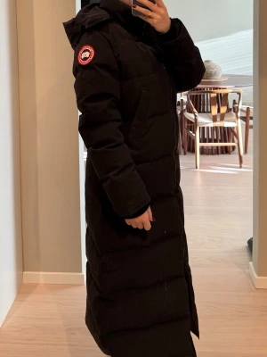 Svart lång dunjacka från Canada Goose - Säljer en svart lång dunjacka från Canada Goose med huva och dragkedja framtill. Jackan har en klassisk pufferlook och patch med logga på ärmen. Perfekt för kalla dagar och riktigt snygg till vintern. Storlek M men passar bra till S/XS
