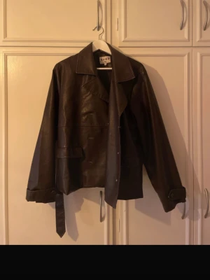 Brun skinnjacka från NA-KD - Snygg brun skinnjacka från NA-KD med klassisk krage, knappar och bälte i midjan. Jackan har långa ärmar och en stilren design som passar perfekt till många olika outfits. Nypris 999kr