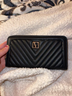 Svart quiltad plånbok från Victoria Secret  - Snygg svart plånbok från VS med quiltad design och guldfärgad logga framtill. Tillverkad i skinn med dragkedja runtom. Perfekt storlek för kort, cash och mynt. Stilren och trendig accessoar som lyfter vilken outfit som helst.