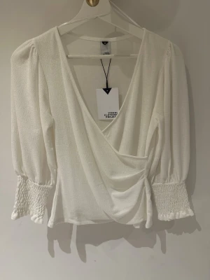 Vit omlottblus från Visual Clothing Project storlek M - Stilren vit omlottblus från Visual Clothing Project med djup V-ringning och puffiga långärmar. Ärmarna har breda smockade muddar och tyget är lätt strukturerat för en trendig look. Perfekt att matcha med både jeans och kjol.