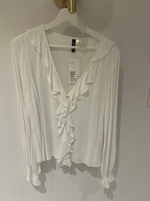 Vit volangblus från H&M storlek 38 - Stilren vit blus från H&M med långa ärmar och volangdetaljer längs den djupa v-ringningen. Tillverkad i ett lätt, krinklat tyg som ger en luftig känsla. Blusen har knapp framtill och smock vid ärmsluten för extra detalj.