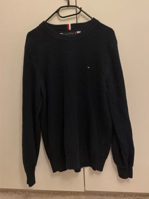 Mörkblå stickad tröja Tommy Hilfiger - Snygg mörkblå stickad tröja från Tommy Hilfiger med diskret logga på bröstet. Tröjan har rund halsringning, ribbade muddar och ett strukturerat stickat mönster. Perfekt för dig som gillar klassisk och stilren design.