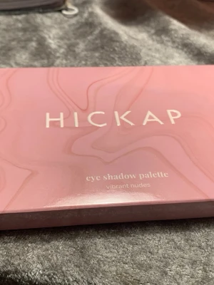 Hickap ögonskuggspalett Vibrant Nudes - Snygg ögonskuggspalett från Hickap med 12 olika nyanser i nude och jordnära toner, både matta och skimrande. Paletten har en rosa förpackning med spegel och färger som beige, guld, orange, brun och grå. Perfekt för kreativa sminkningar och naturliga looks.
