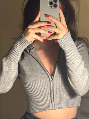 Grå croppad hoodie med dragkedja - Snygg grå croppad hoodie med dragkedja framtill och ribbade detaljer vid midja och ärmslut. Hoodien har en huva och är tillverkad i ett mjukt stickat material som ger en skön och trendig look. Perfekt för en avslappnad och stilren outfit.