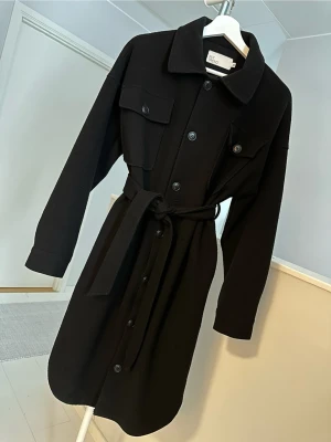 Svart long shacket med bälte från Nelly - Stilren svart schaket med klassisk krage, knappar längs hela framsidan och två bröstfickor. Jackan har ett matchande bälte i midjan och långa ärmar. Oversize, är 32 men mer som en 34/36. 