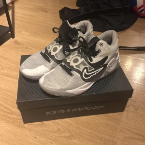 Nike KD Trey 5X grå sneakers - Snygga Nike KD Trey 5X sneakers i grått och svart med coola detaljer och Kevin Durant-logga på sidan. Skorna har snörning och är tillverkade i syntetmaterial och mesh för bra andningsförmåga. Perfekta för dig som gillar basketstil och streetwear.