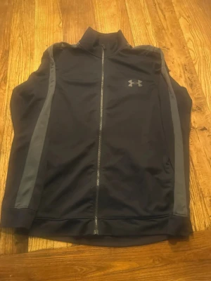 Svart träningsset från Under Armour - Snyggt svart träningsset från Under Armour med gråa detaljer längs sidorna. Setet består av en zip-jacka med hög krage och matchande byxor med dragsko i midjan. Perfekt för gymmet eller löprundan. Materialet är mjukt och stretchigt för maximal rörelsefrihet.