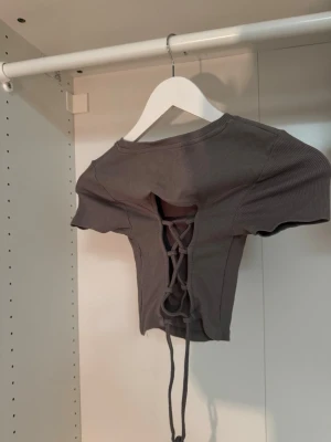 Grå croppad topp med snörning - Snygg grå croppad topp med korta ärmar och ribbad struktur. Toppen har en cool snörningsdetalj i ryggen som ger en edgy vibe. Perfekt att styla med höga jeans eller kjol för en trendig look🤍