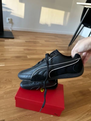 Svarta Puma Ferrari sneakers - Snygga svarta Puma Ferrari sneakers med klassisk vit logga på sidan och detaljer i svart skinn. Skorna har snörning och en låg profil, samt Ferrari-märke på plösen. Perfekta för dig som gillar sportig stil och exklusiva samarbeten.