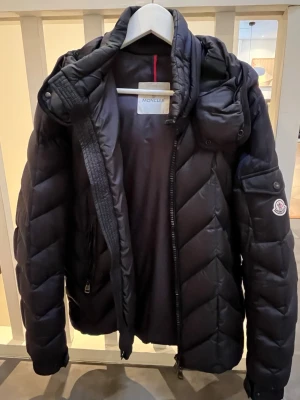 Svart Moncler dunjacka med huva - Säljer en svart Moncler dunjacka med huva och dragkedja. Jackan har quiltad design, Moncler-logga på ärmen och praktiska fickor. Perfekt för kalla dagar och riktigt snygg till vintern.