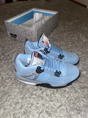 Nike Air Jordan 4 University Blue - Snygga Nike Air Jordan 4 University Blue sneakers i ljusblå mocka med svarta och grå detaljer. Skorna har snörning, meshpaneler och en vit sula med svart och grått stänkmönster. Perfekta för dig som vill sticka ut med klassisk Jordan-stil.