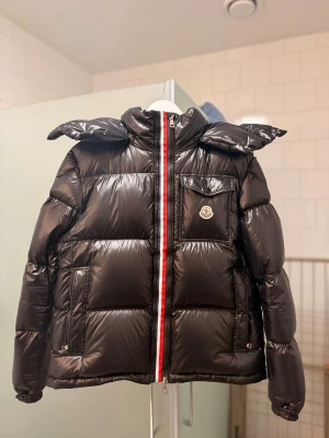 Svart dunjacka från Moncler - Svart glansig dunjacka från Moncler med hög krage och ikonisk logotyp på bröstfickan. Jackan har quiltad design, dragkedja framtill och en rödvit rand längs dragkedjan på ena bilden. Perfekt för kalla dagar.