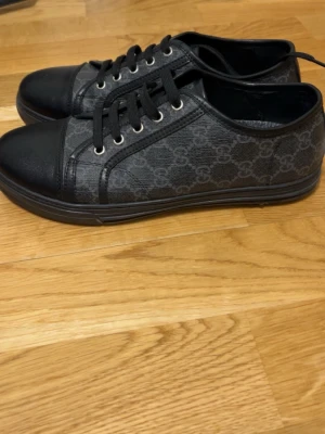 Svarta sneakers med GG-mönster - Stilrena svarta sneakers från Gucci med klassiskt GG-mönster på sidan. Skorna har svarta snören, rund tå och detaljer i skinn. Perfekta för dig som gillar en clean och lyxig look.