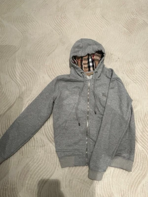 Burberry kofta - Burberry kofta i grått – storlek S Stilren och tidlös kofta med dragkedja. Mjuk och bekväm med Burberrys klassiska rutmönster i huvan som ger en exklusiv känsla. I fint skick och lätt att matcha till alla tillfällen