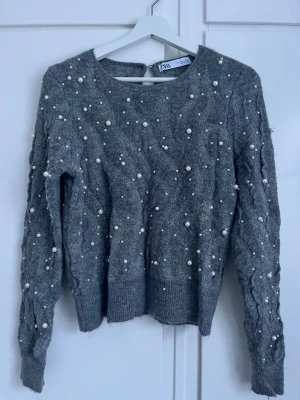 Grå stickad tröja med pärlor från ZARA - Trendig grå stickad tröja från ZARA med dekorativa vita pärlor över hela plagget. Tröjan har rund halsringning, kabelstickade detaljer på ärmarna och ribbade muddar vid ärmslut och nederkant. Perfekt för dig som vill ha något extra till din outfit.