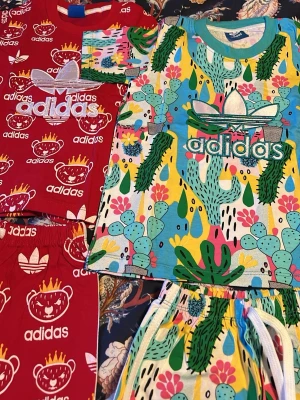 Färgglada Adidas tröjor med matchande byxor i tunt material - Två coola Adidas tröjor med byxor, en röd med vita loggor och tecknade ansikten med kronor, och en färgglad med kaktusar, blommor och stora Adidas-tryck. Båda har korta ärmar och är perfekta för dig som gillar färg och mönster. Står L i kläderna men skulle säga det är som barnstorleken 86