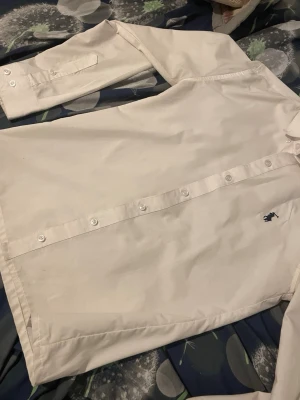 Vit skjorta från U.S. Polo Assn - Klassisk vit skjorta från U.S. Polo Assn med lång ärm och knappar framtill. Skjortan har en broderad logga i mörkblått på bröstet och är tillverkad i bomull. Perfekt till en clean och stilren look.