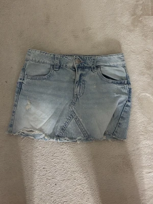 Ljusblå jeanskjol med rå kant - Trendig ljusblå jeanskjol i minilängd med rå, fransig nederkant och slitna detaljer. Kjolen har klassiska fickor fram och bak samt en snygg sömdetalj framtill. Perfekt för dig som gillar en avslappnad och cool stil.