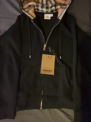 Svart hoodie med dragkedja från Burberry - Snygg svart hoodie från Burberry med dragkedja och klassiskt rutigt foder i huvan. Hoodien har justerbara snören och ribbade muddar. Tillverkad i mjukt bomullsmaterial och har en exklusiv känsla med Burberry-tag framtill.