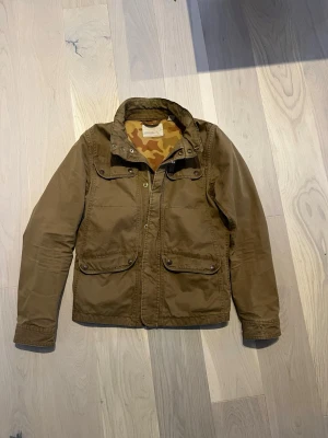 Scotch & soda armyjacka - Snygg brun army jacka med camouflagemönstrat innerfoder och praktiska fickor framtill. Jackan har hög krage, långa ärmar och stängs med dragkedja och knappar. Perfekt för dig som vill ha en stilren och funktionell jacka till vardags.