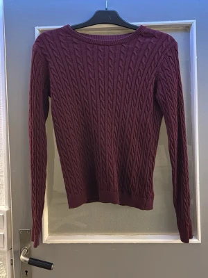 Vinröd kabelstickad tröja - Snygg vinröd kabelstickad tröja med rund hals och långa ärmar. Tröjan har ribbade muddar vid ärmslut och nederkant, vilket ger en klassisk och stilren look. Perfekt för dig som gillar en enkel men trendig stil.