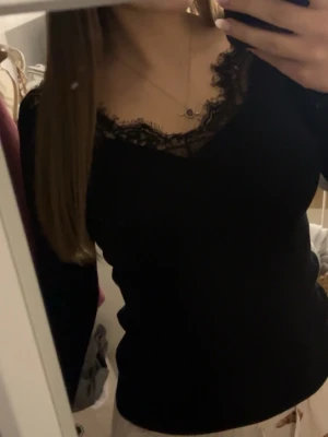 Svart ribbad topp med spetsdetalj - Snygg svart långärmad topp med ribbad struktur och v-ringning dekorerad med spets. Perfekt att styla med jeans eller kjol för en trendig look. Figurnära passform och mjukt material som känns skönt mot huden.